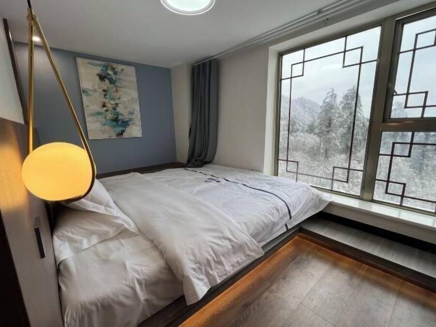 Un dormitorio con cama, ventana con vista a árboles, un cuadro en la pared y una lámpara.