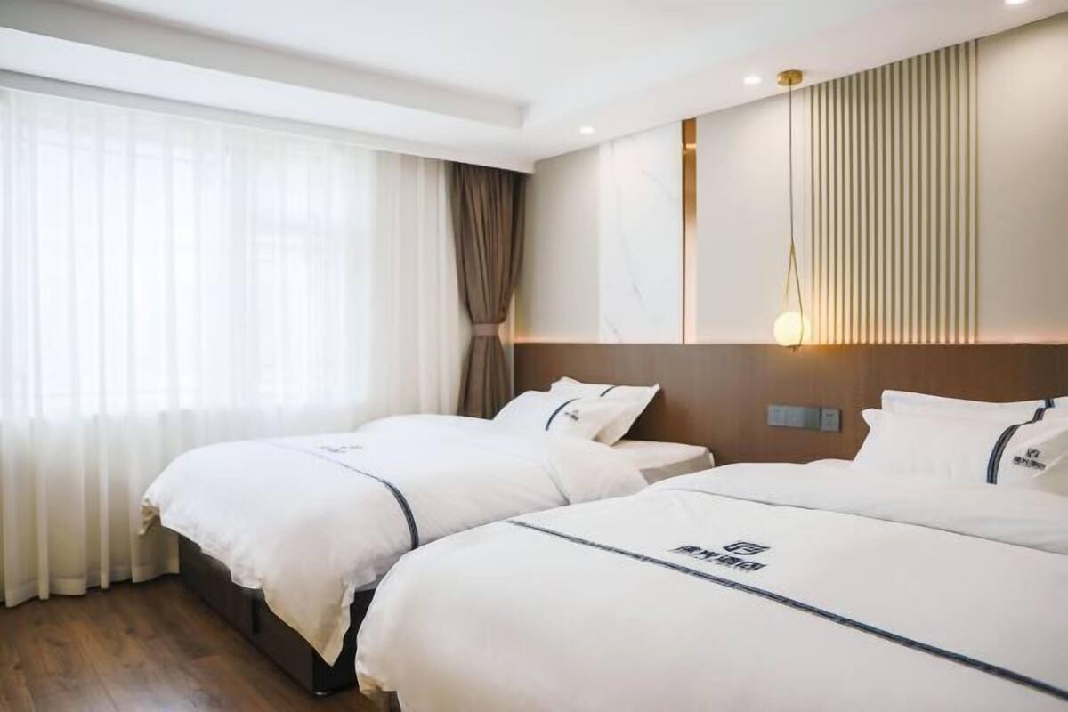 A hotel room with two beds, a large window with sheer curtains, a wall-mounted light, and a wooden floor.