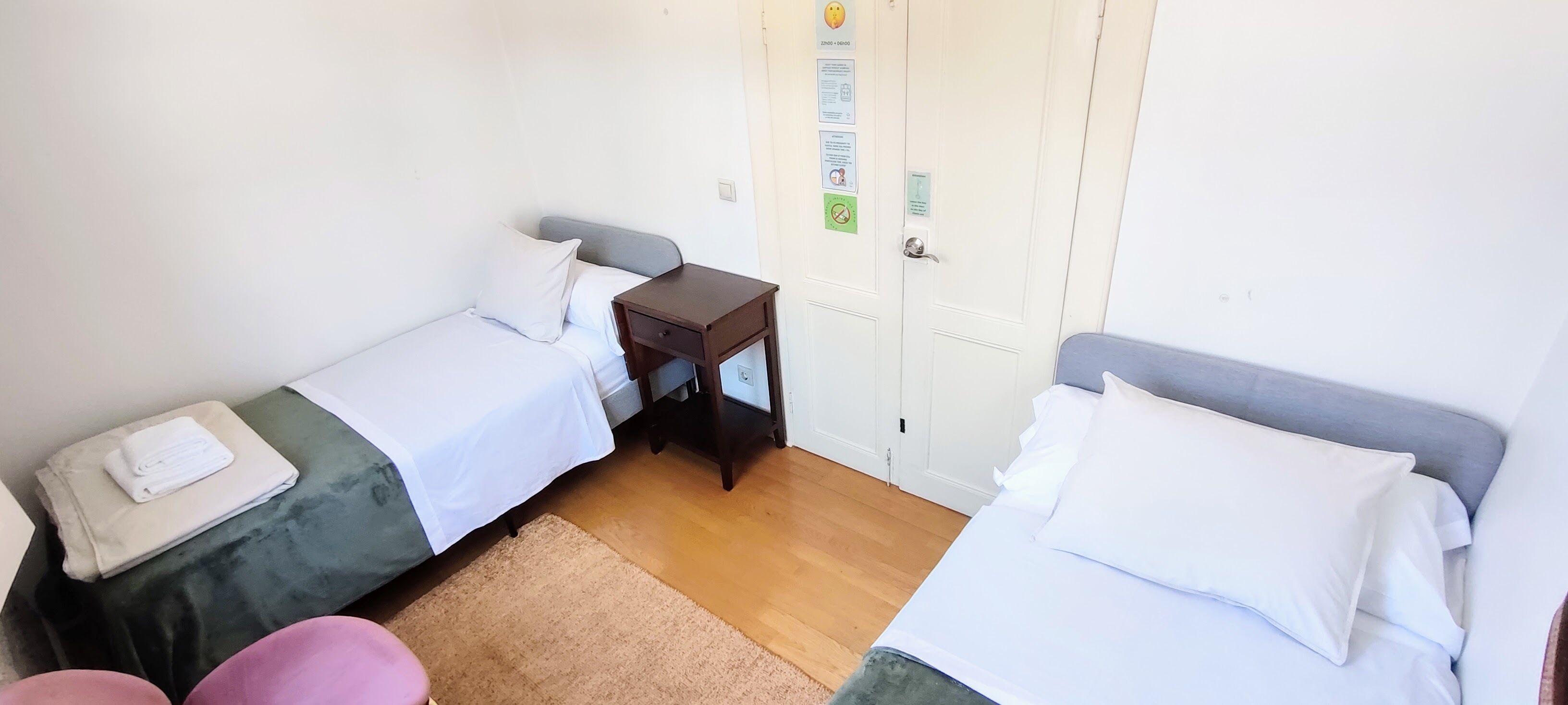 Una habitación pequeña y limpia con dos camas individuales, una mesita de noche de madera, una pequeña mesa y una puerta con cerradura.