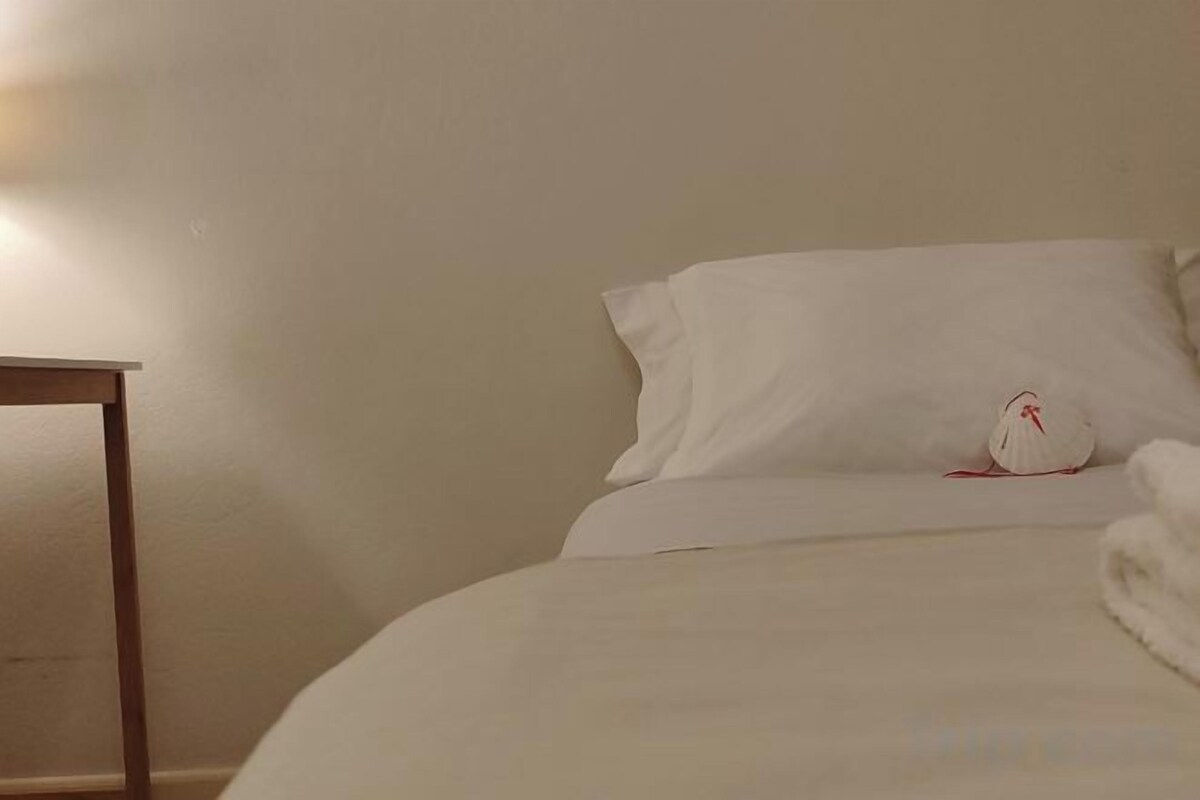 A neatly made bed with white linens and pillows, a wooden nightstand, and a wall-mounted light.