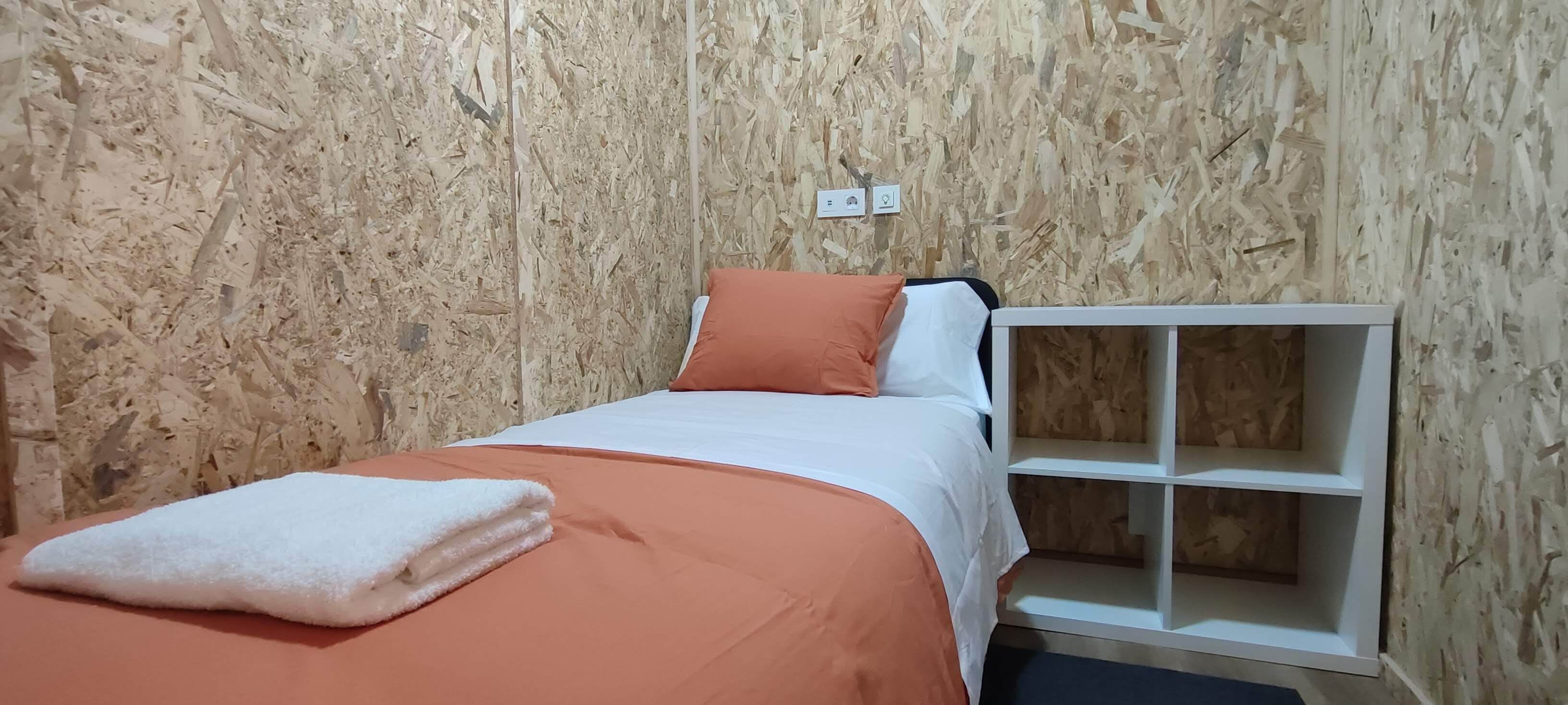 Un llit individual amb un estovat taronja i almoines blanques, una taula de nit blanca i un prestatge blanc al costat d'una paret de fusta.