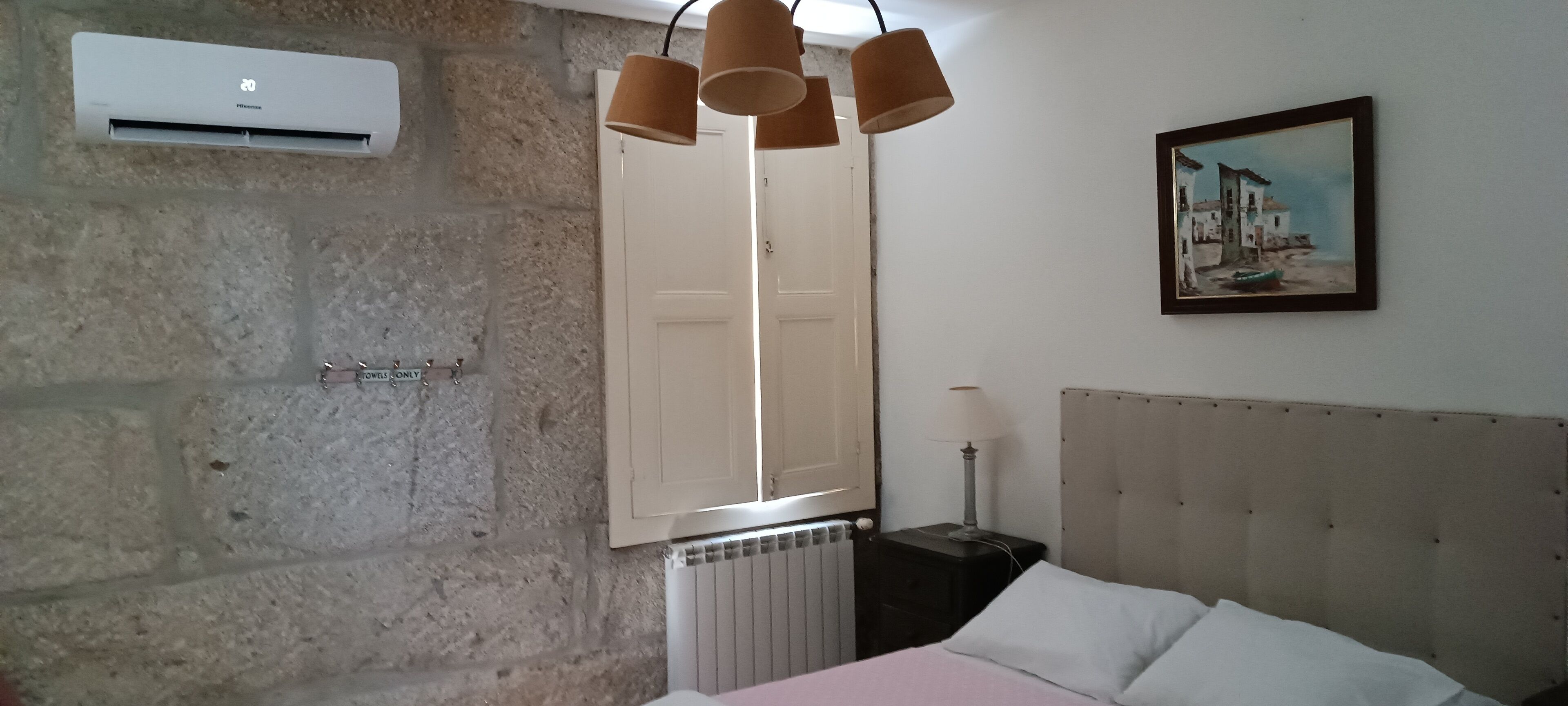 Un dormitorio con pared de piedra, una cama con cabecera capitonada, un cuadro, un radiador y una lámpara colgante.