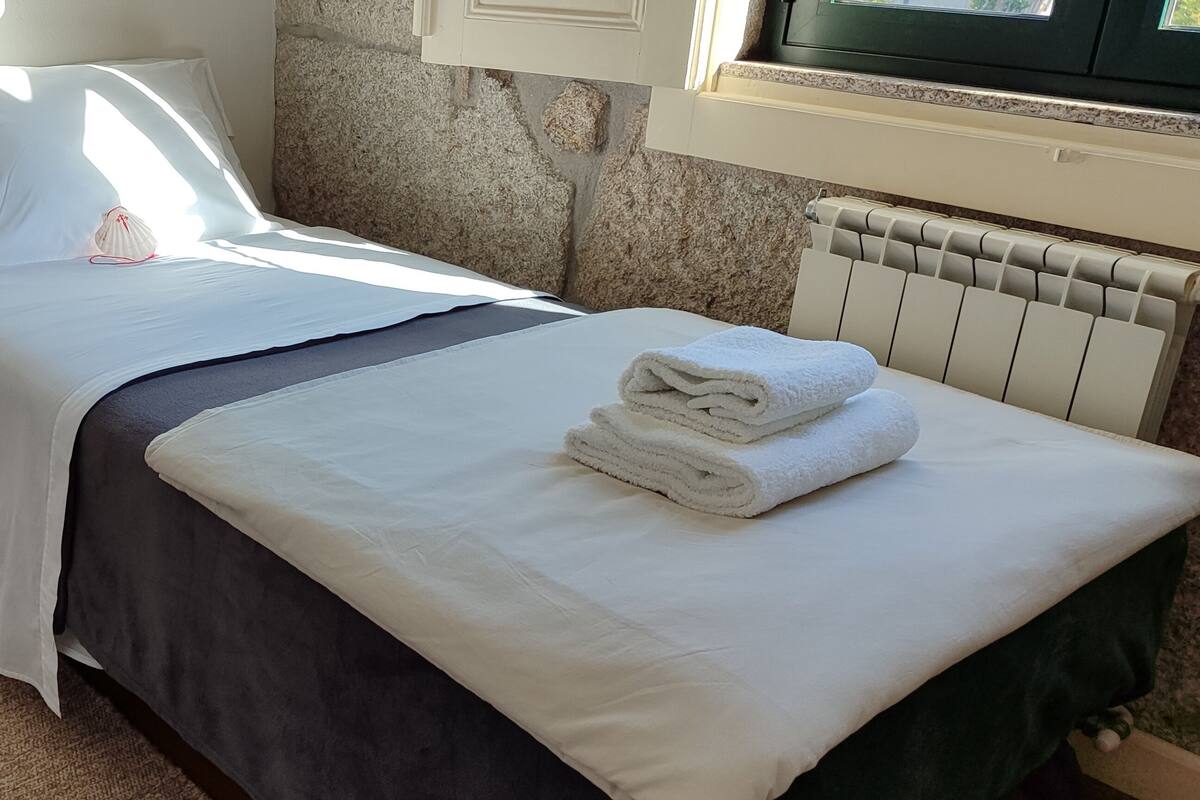A neatly made bed with white linens and a folded towel, a stone wall, and a radiator.