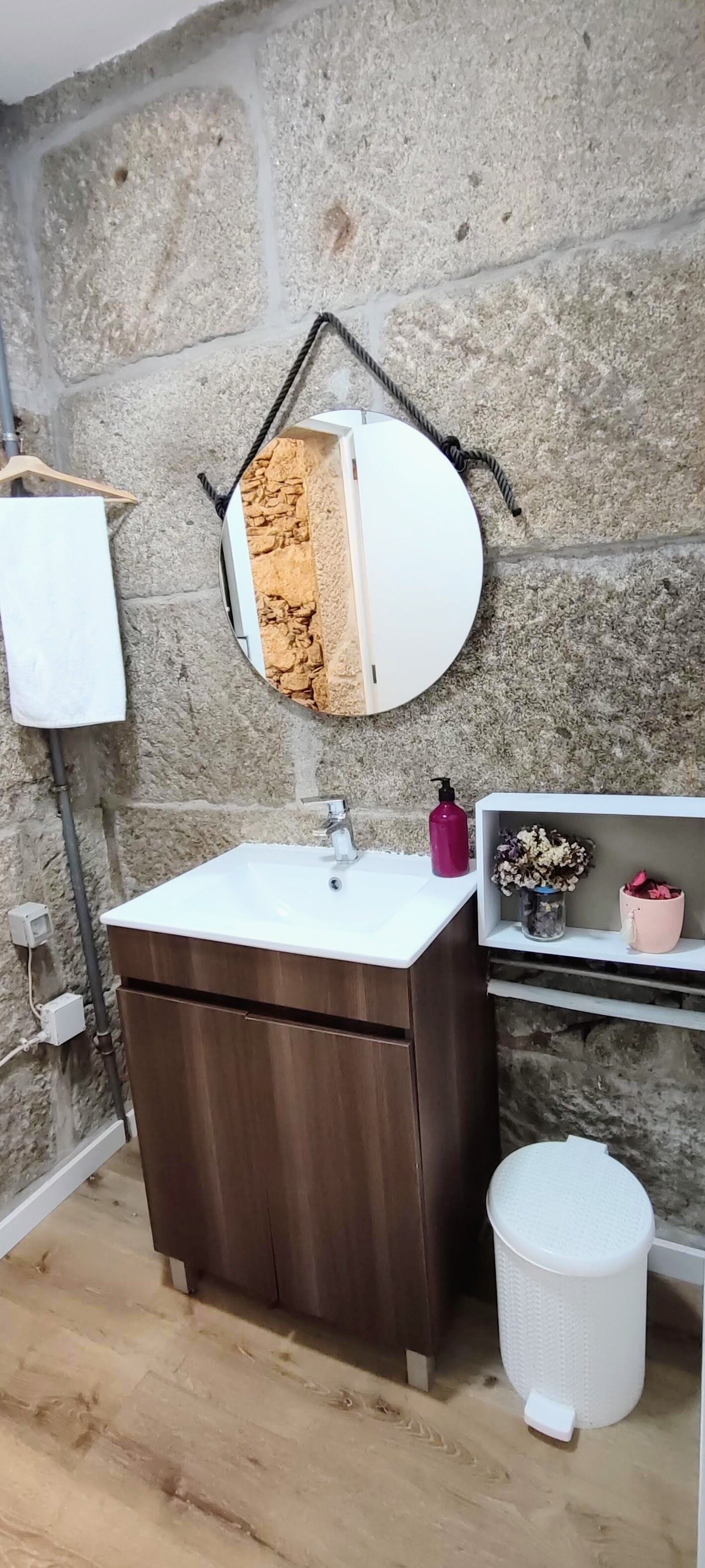A bathroom with a stone wall, a wooden vanity, a white sink, a mirror, a hanging towel, a shelf with toiletries, and a trash bin.
