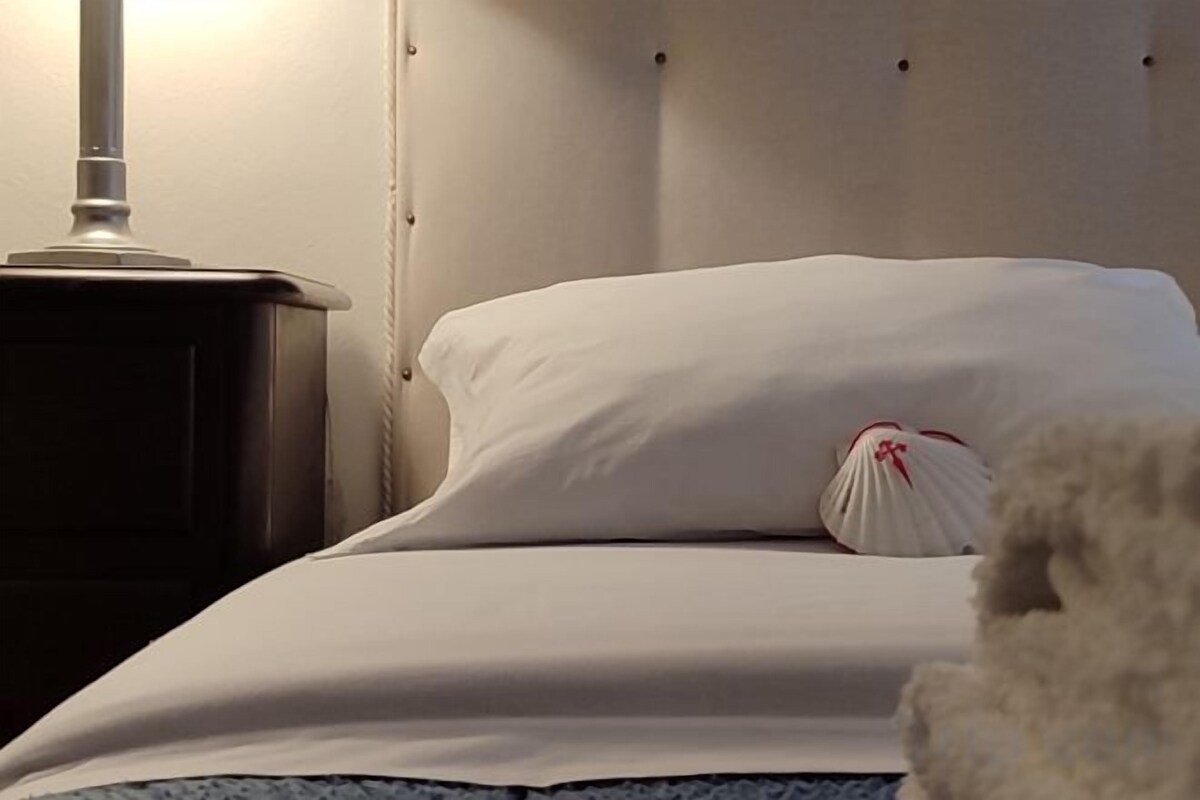 A bed with white bedding and a pillow with a red design, a bedside table with a lamp, and a fluffy white object on the bed.