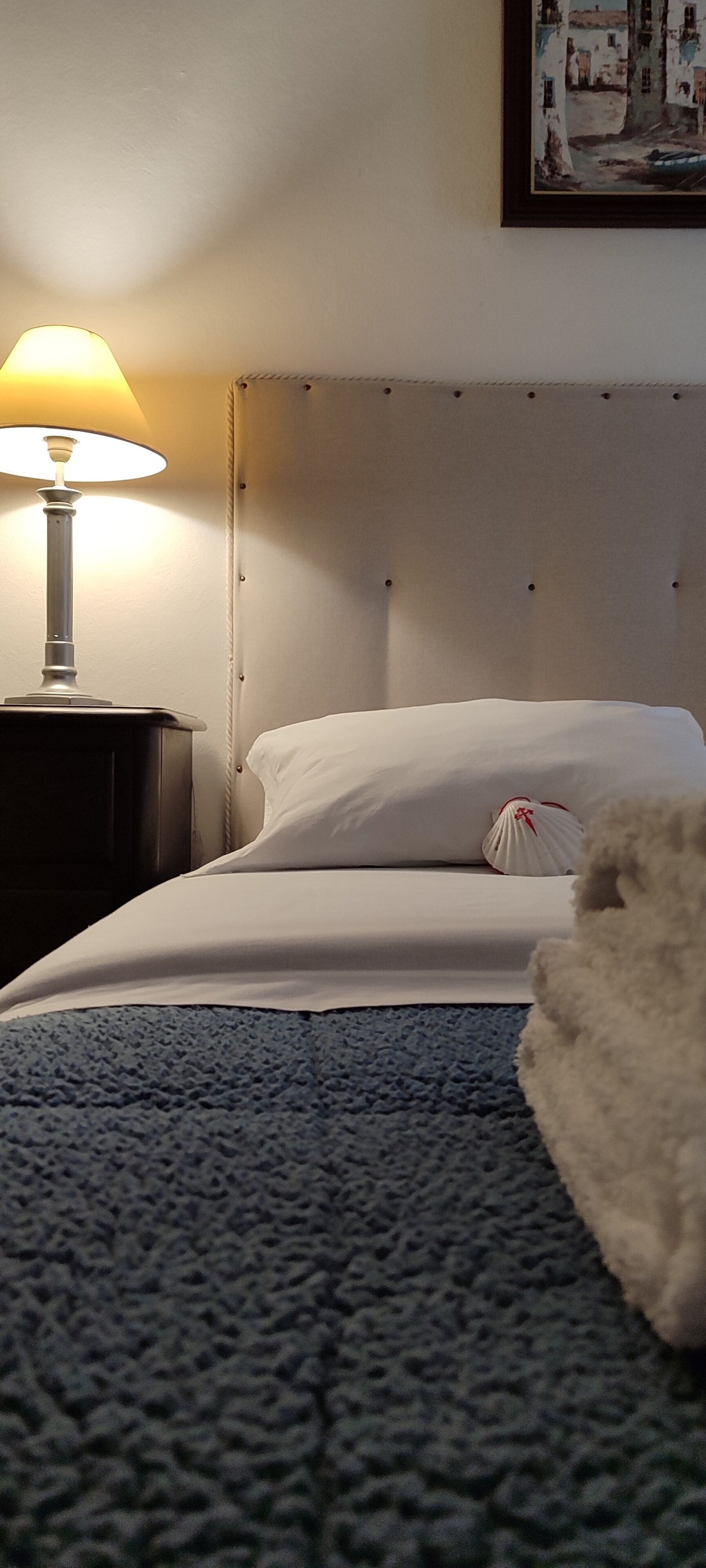Un lit bien fait, avec des draps blancs, une couverture bleue à texture et un oreiller blanc et moelleux. Une lampe avec un abat-jour blanc est posée sur une table de chevet en bois foncé, à côté du lit.