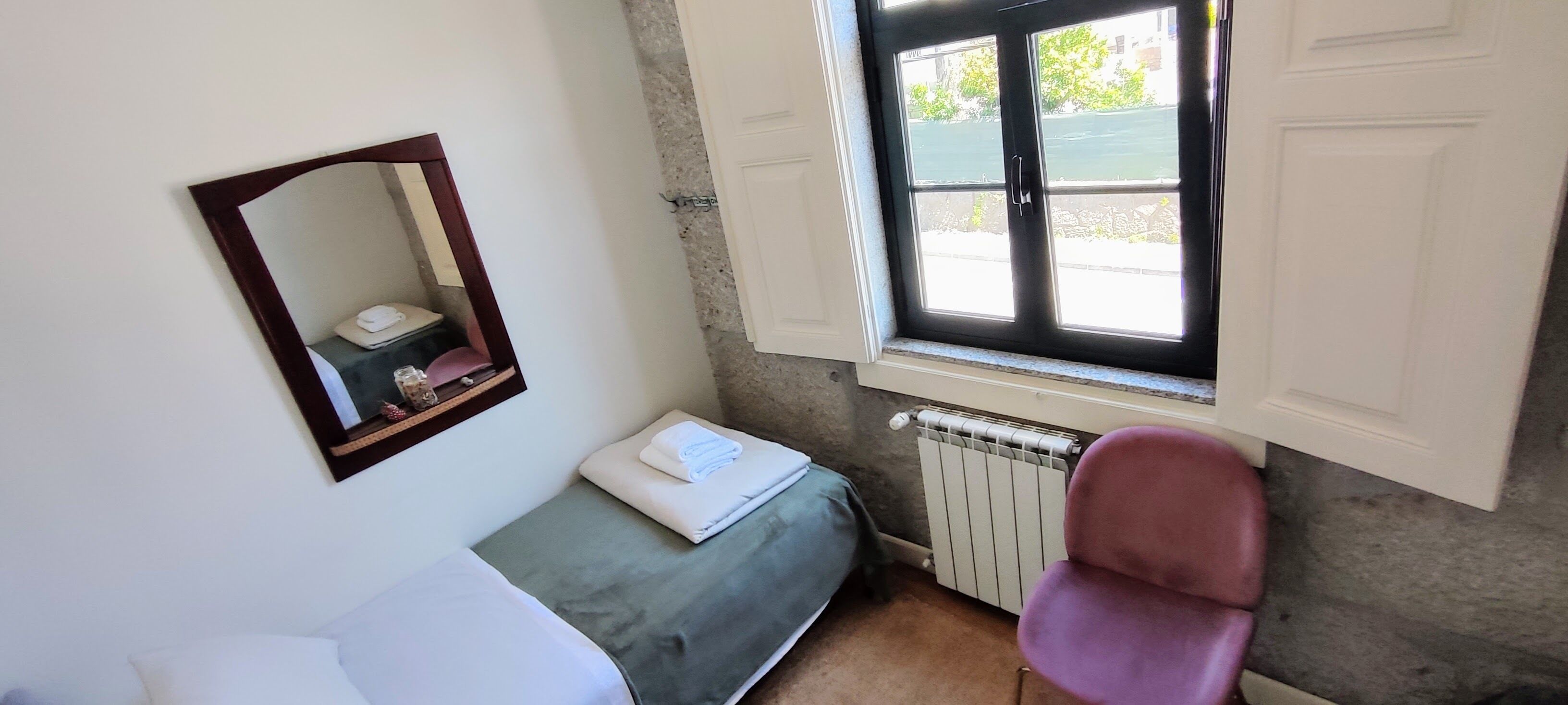 A small, compact room with a single bed, a chair, a radiator, and a window with a view of the outdoors.