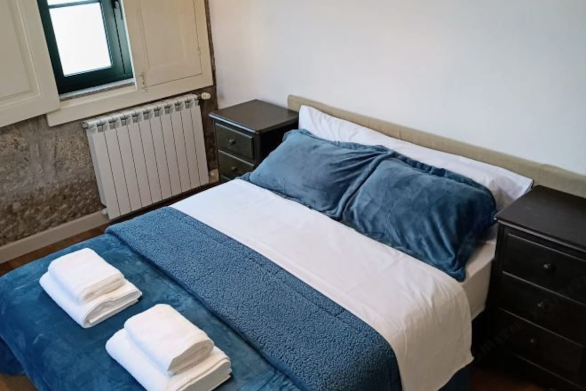 A neatly made bed with blue and white bedding, a small bedside table, a radiator, and a window with blinds.