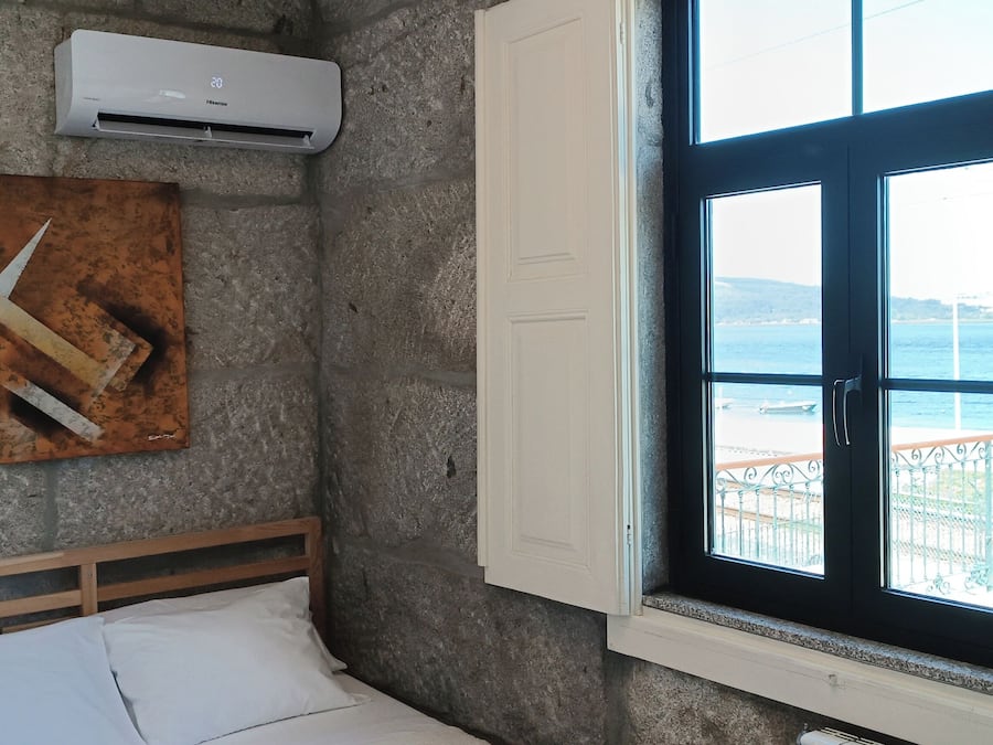 A bedroom with a stone wall, a bed with white linens, a painting, an air conditioning unit, and a window with a view of the sea.