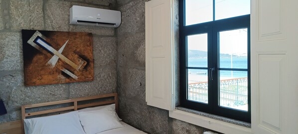 A bedroom with a stone wall, a bed with white linens, a painting, an air conditioning unit, and a window with a view of the sea. - Donna Nega Guest House  (Caminha)