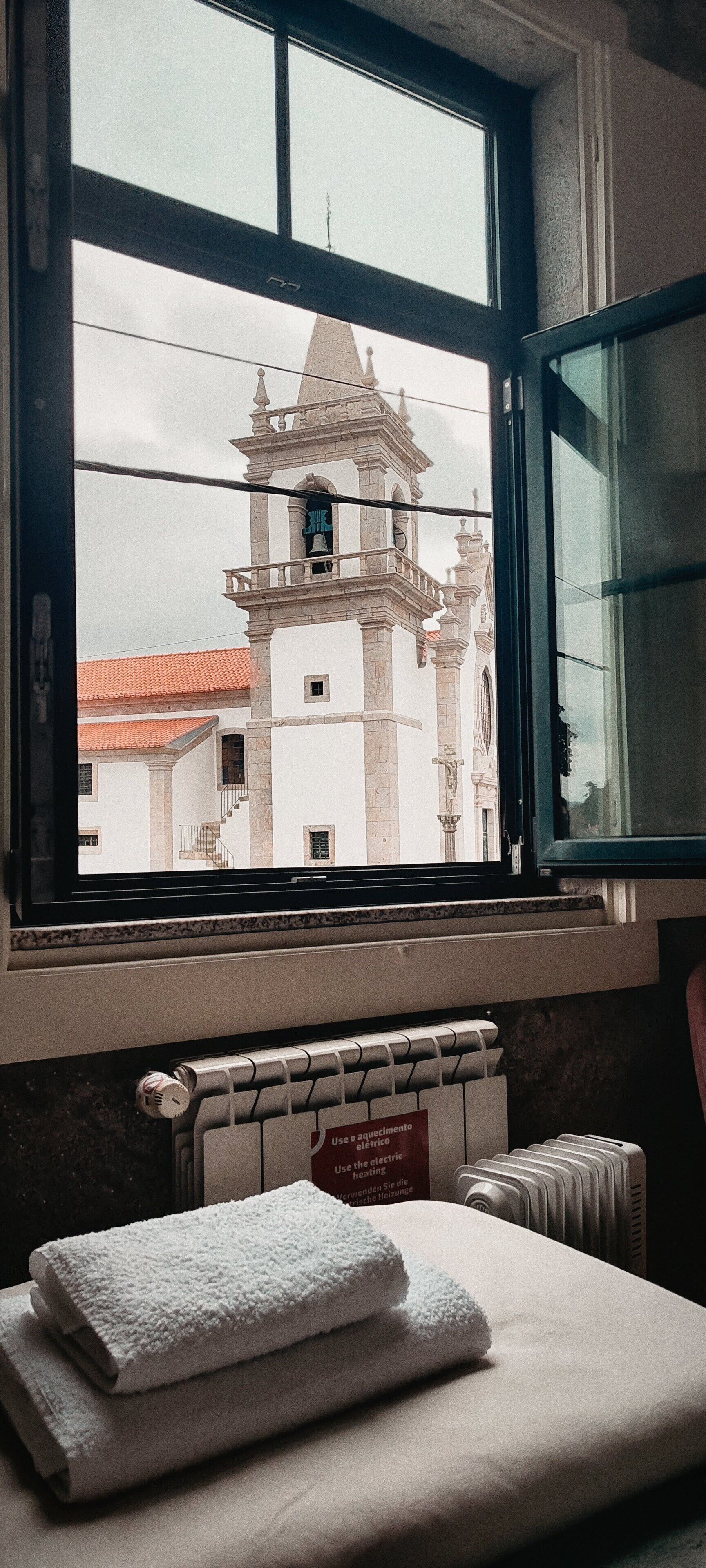A bathroom with a window overlooking a building with a bell tower. There is a towel on the counter and a radiator below the window.