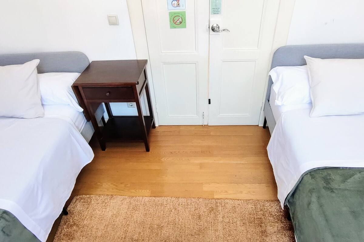 A room with two single beds, a small wooden table, and a door with a green sign.