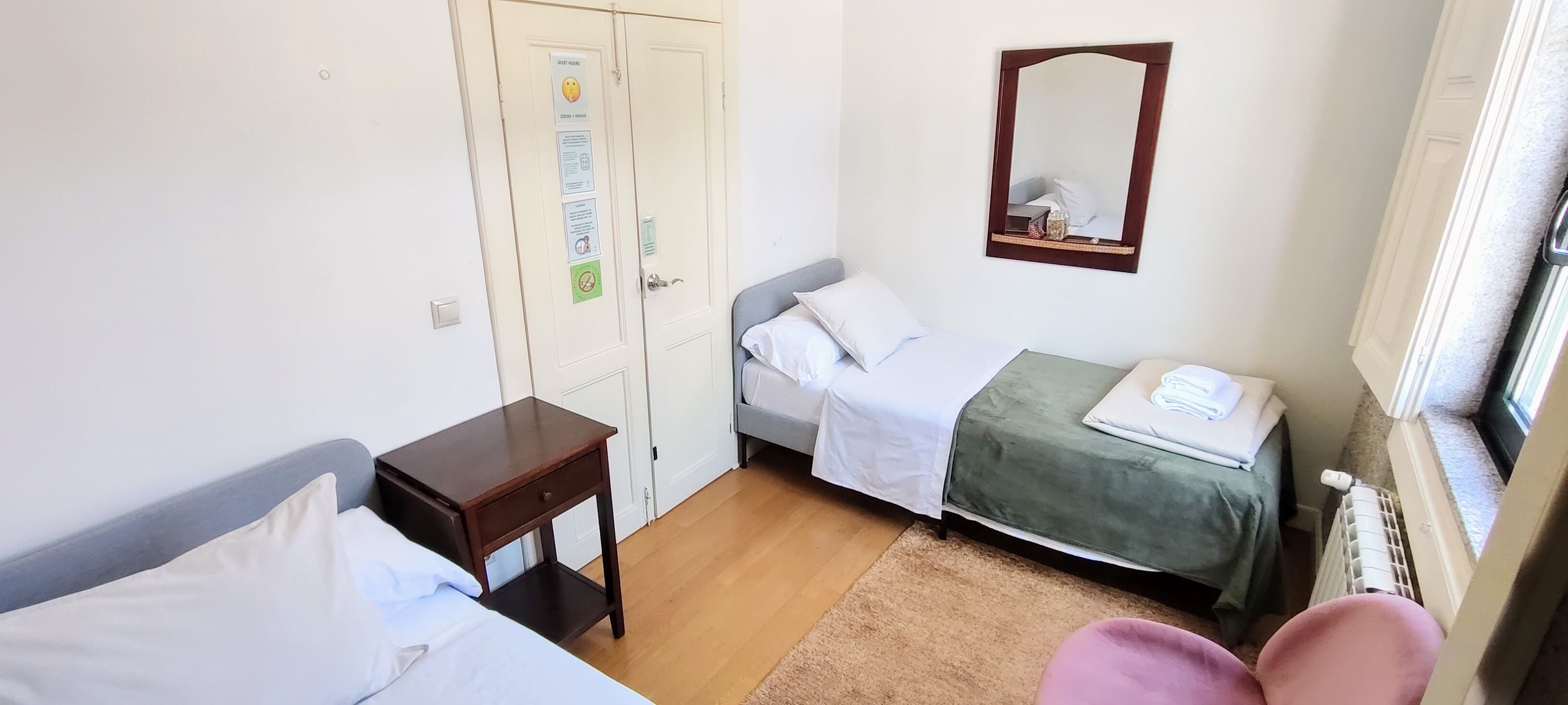 A small, clean room with two beds, a wooden nightstand, a mirror, and a window with a view.