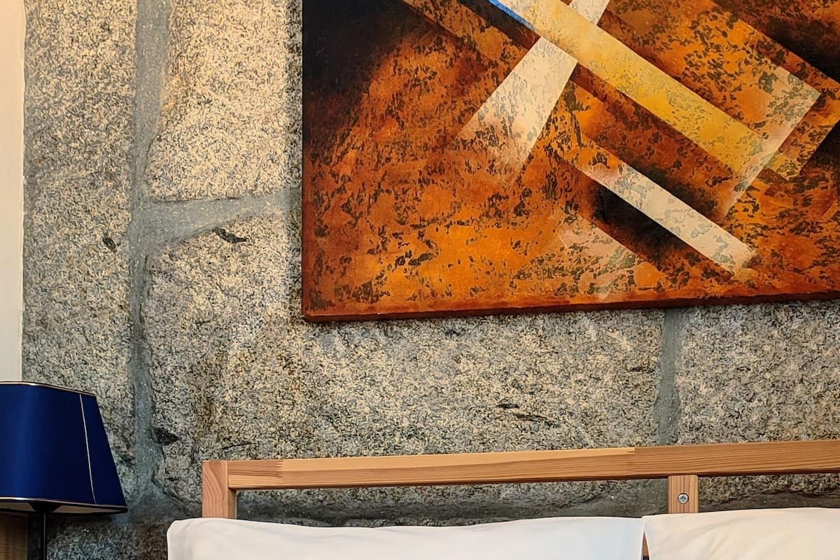 A bed with white and blue bedding, a wooden headboard, and a stone wall with a painting.