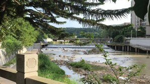 Um rio com uma ponte e uma cachoeira, rodeado por vegetação e edifícios ao fundo.