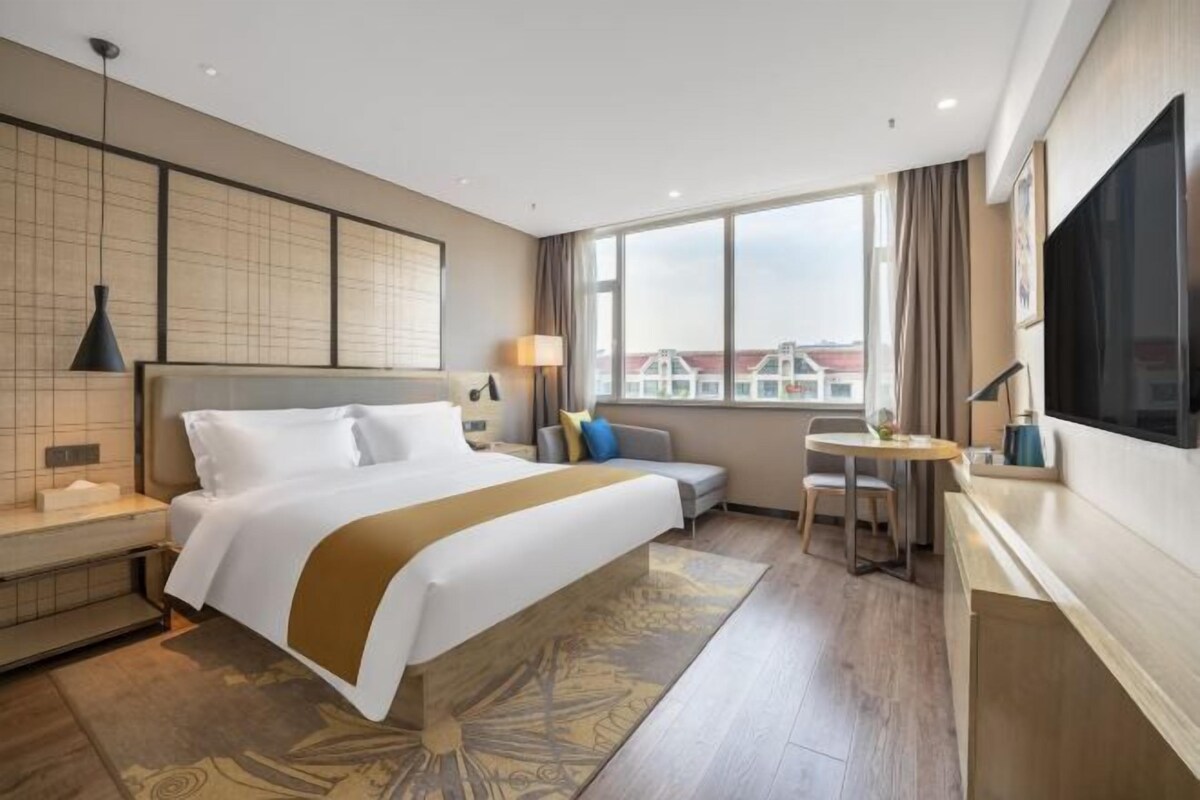 A hotel room with a large bed, a desk, a chair, a TV, and a view of buildings outside the window.