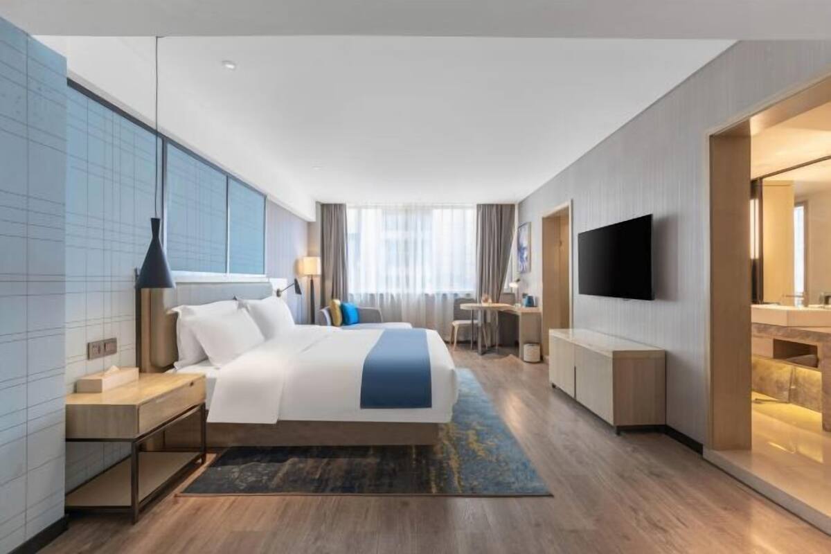 A modern hotel room with a large bed, bedside tables, a desk, a chair, a TV, and a bathroom with a mirror and sink.