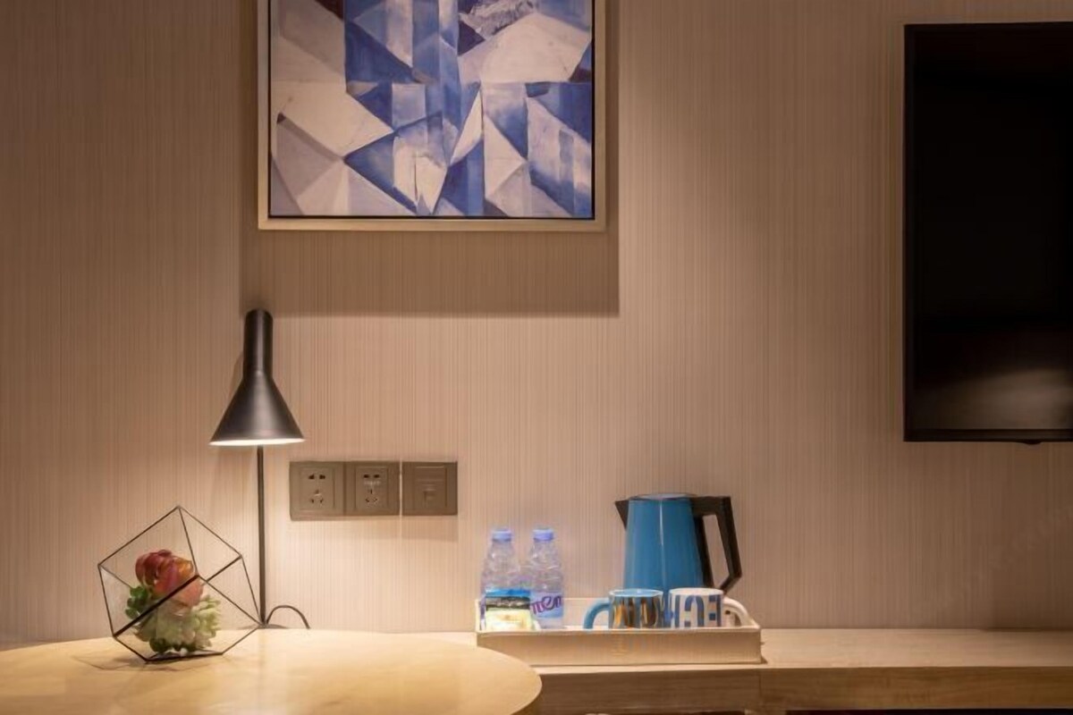 A hotel room with a wooden table, a geometric vase, a lamp, a coffee maker, and a flat-screen TV.