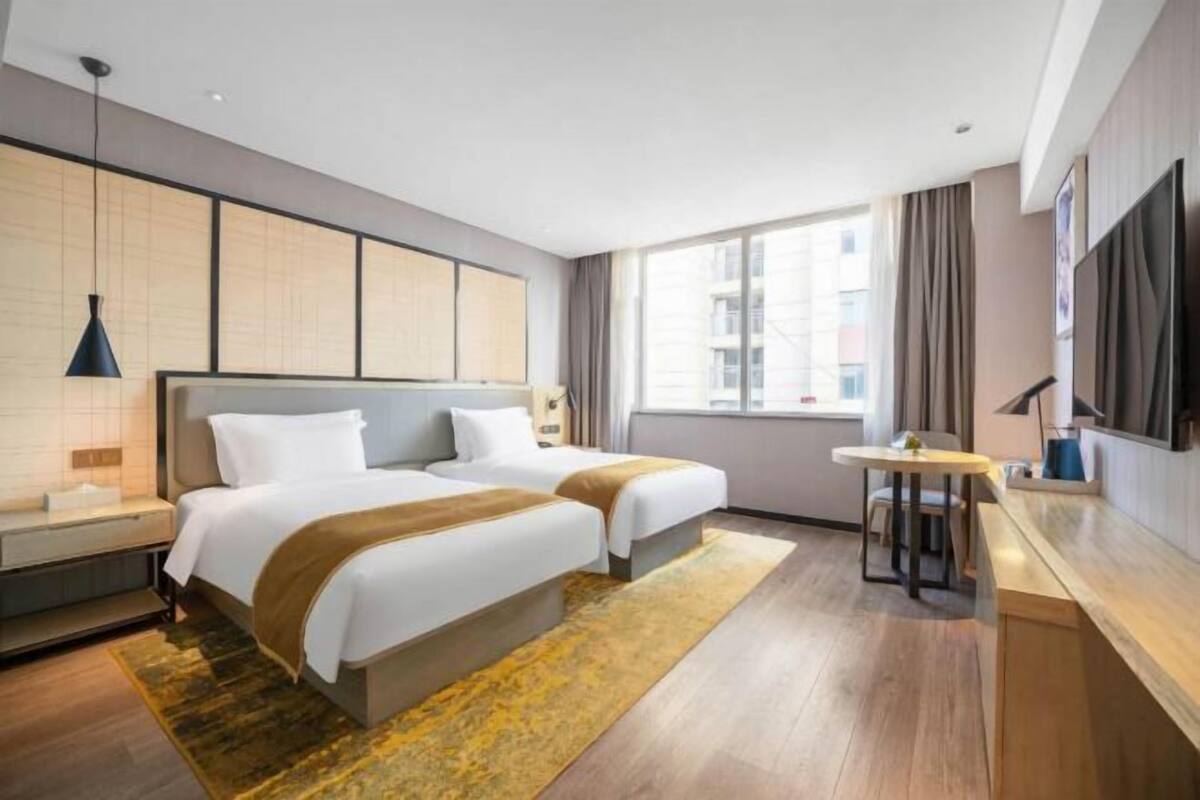 A hotel room with two beds, a small table, a TV, and a window with a view of buildings.