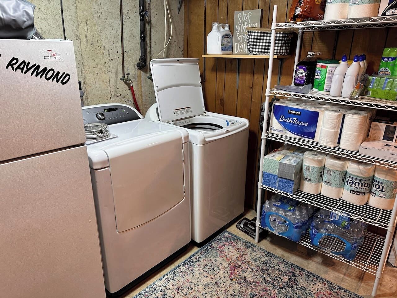 A laundry room with a Raymond brand washer and dryer, a shelf with bath products, and a rack with toilet paper and paper towels.