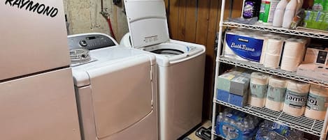 A laundry room with a Raymond brand washer and dryer, a shelf with bath products, and a rack with toilet paper and paper towels.