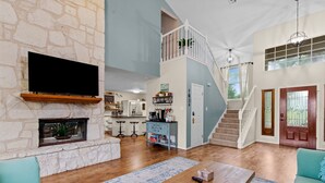 A spacious living area with a stone fireplace, a kitchen in the background, a staircase leading to an upper level, and a large window with a view. - Hot Tub/Shuffleboard/Pool Table near Guadalupe Riv. & Lake (New Braunfels)