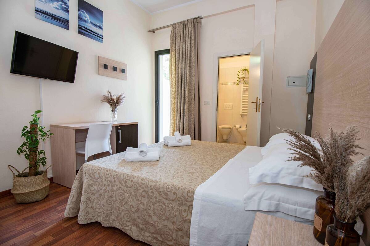 Une chambre d’hôtel avec un lit, un bureau et une chaise, un téléviseur fixé au mur, et une salle de bain visible à travers une porte ouverte.