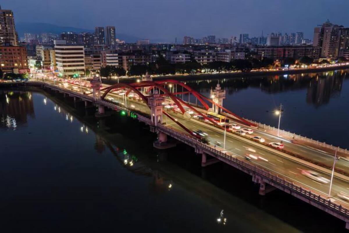 A nighttime cityscape with a prominent red bridge over a river, surrounded by buildings and a calm water surface reflecting city lights.