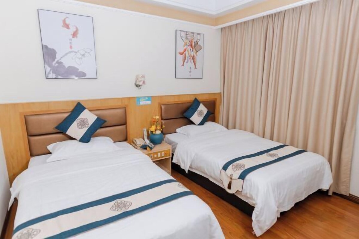A hotel room with two beds, each with a blue-striped bedspread and matching pillows. There are framed artworks on the walls and a curtain on the window.