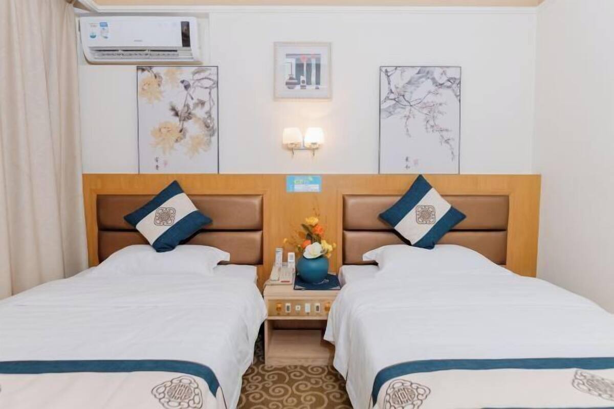 A hotel room with two beds, a nightstand with a vase of flowers, wall-mounted artwork, and an air conditioning unit.