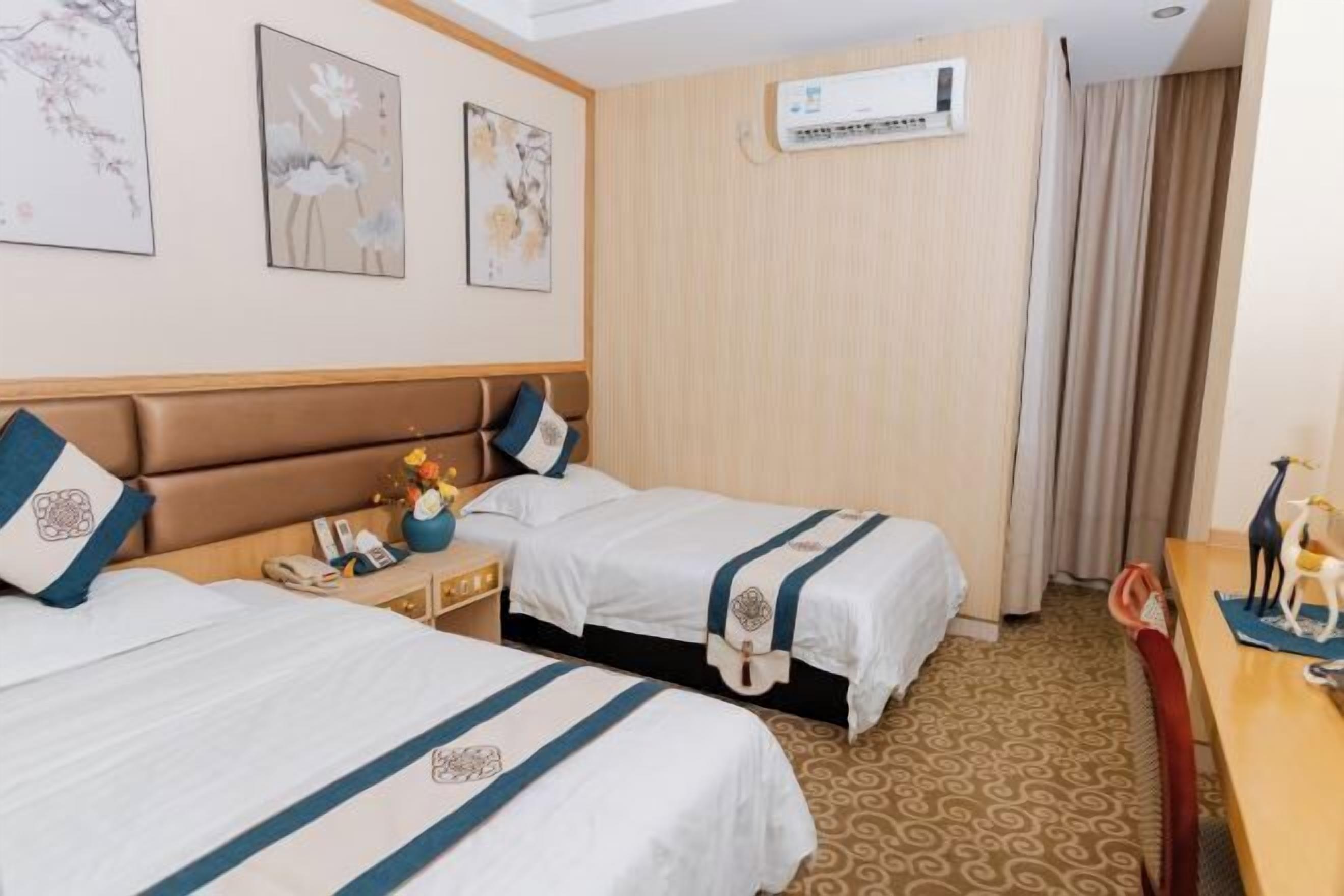 A hotel room with two beds, a wooden headboard, a small table with a vase of flowers, and a wall-mounted air conditioner.