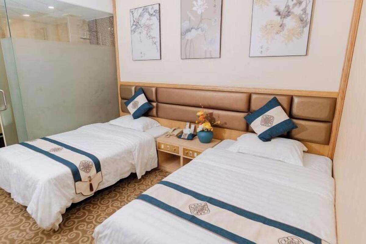 A hotel room with two beds, a wooden headboard, a bedside table with a phone and a vase, and three framed artworks on the wall.