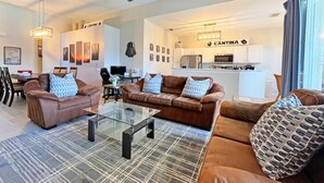 Uma sala de estar moderna com uma mesa de centro de vidro, dois sofás de couro marrom com almofadas estampadas, um ventilador de teto e uma área de cozinha ao fundo.