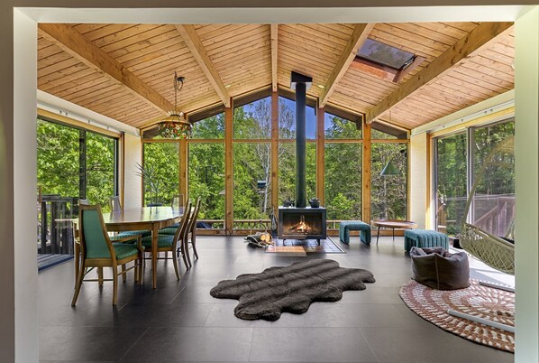 Un espacio amplio con un techo de madera grande, una chimenea, una zona de comedor con sillas y ventanas grandes que ofrecen vistas al exterior.