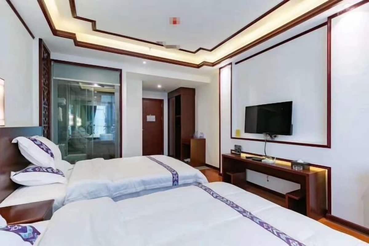 A hotel room with two beds, a TV, a desk, and a glass partition.