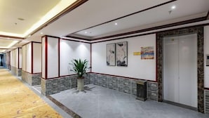 A hotel lobby with a long corridor, a reception desk, a potted plant, artwork on the wall, and an elevator. - Lijin Hotel (Lingshan)