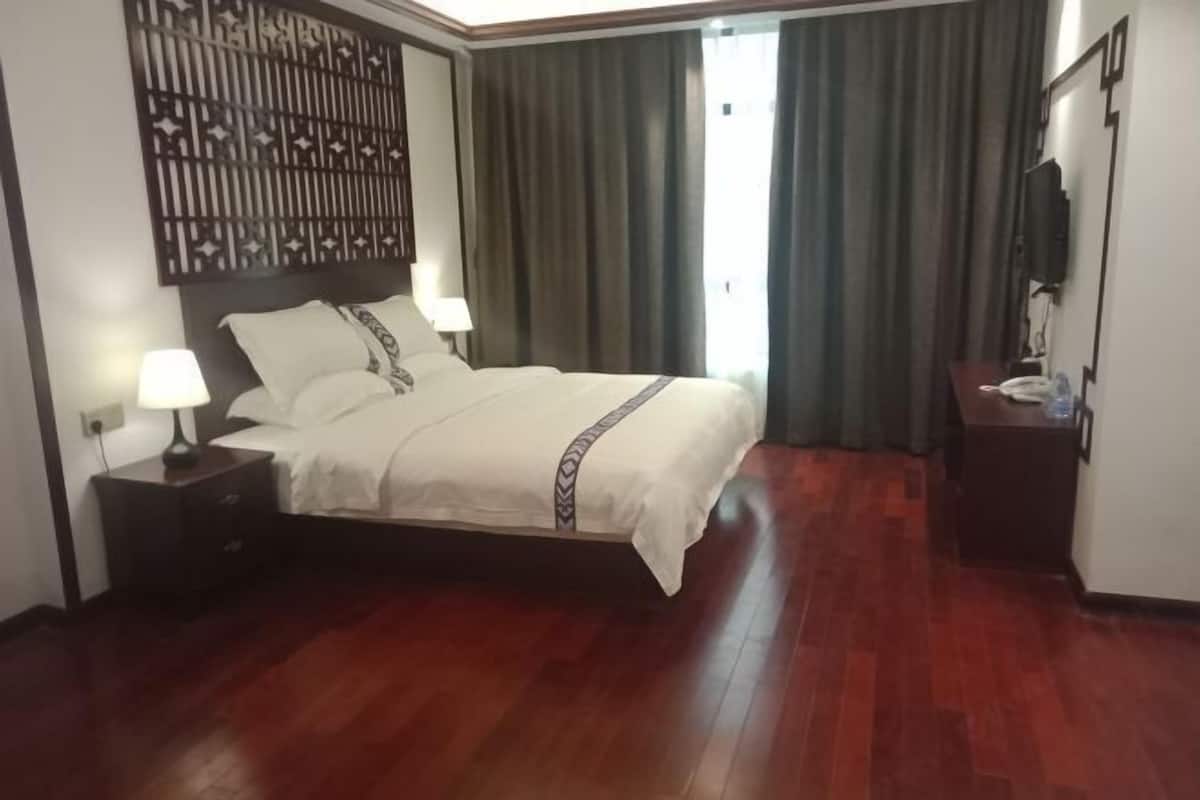 A hotel room with a large bed, a nightstand with a lamp, a TV on a stand, and a window with curtains.