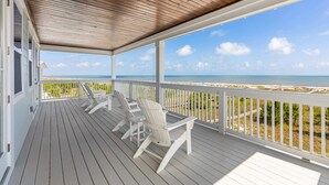 Varanda coberta com grades brancas e cadeiras de deck, com vista para uma praia e céu claro.