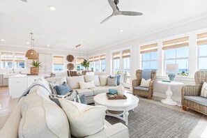 A spacious living room with a large sofa, a coffee table, and wicker chairs. There is a kitchen area with white cabinets and a dining table.