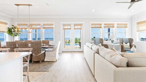 A spacious living area with a large sofa, wicker chairs, a dining table, and a view of the ocean through large windows. - Monterey Oceanfront Beach House (Port St. Joe)