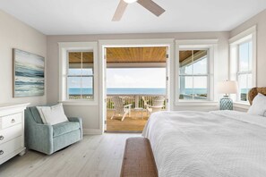 Une chambre avec un grand lit, une table de chevet, une chaise, une fenêtre donnant sur la plage et un ventilateur de plafond.