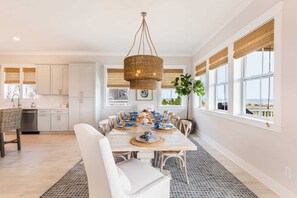 Un coin repas avec une grande table dressée pour six personnes, entourée de chaises blanches, un luminaire suspendu en fibres tressées et une vue sur l’océan grâce à de grandes fenêtres équipées de stores en bambou.