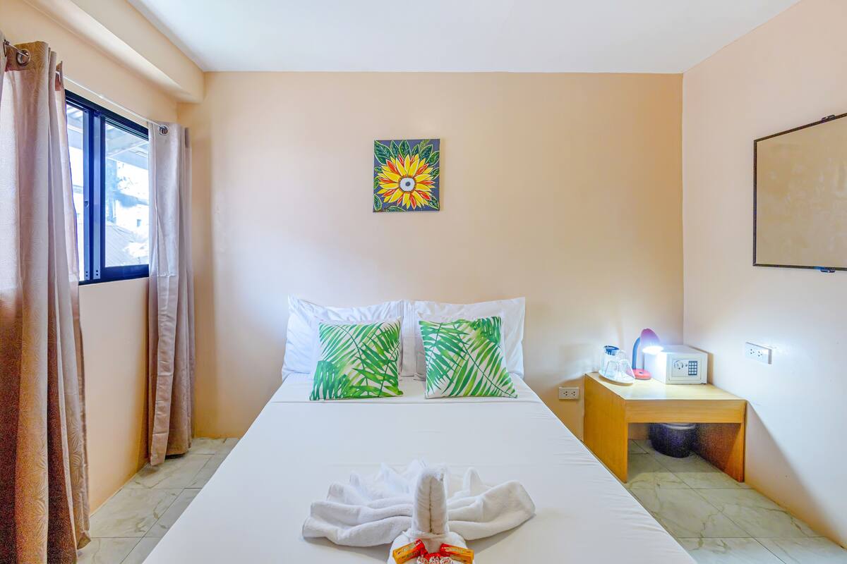 A hotel room with a bed, two pillows, a painting, a bedside table with a lamp, and a window with curtains.