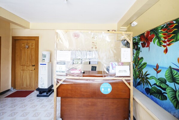 Habitación de hotel con puerta de madera, suelo de baldosas, un cuadro de flores tropicales y un cartel solicitando a los huéspedes mantener la distancia.