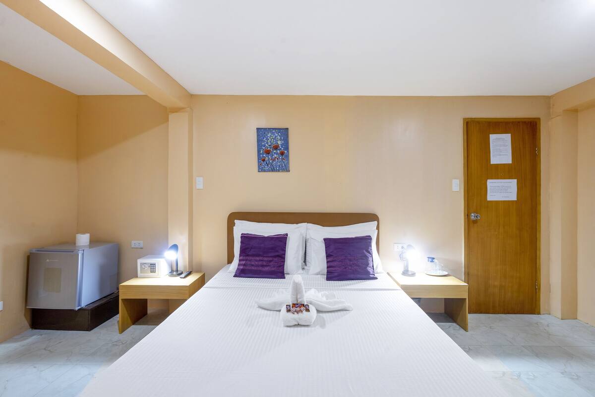 A hotel room with a bed, two nightstands, a wooden door, and a small refrigerator.