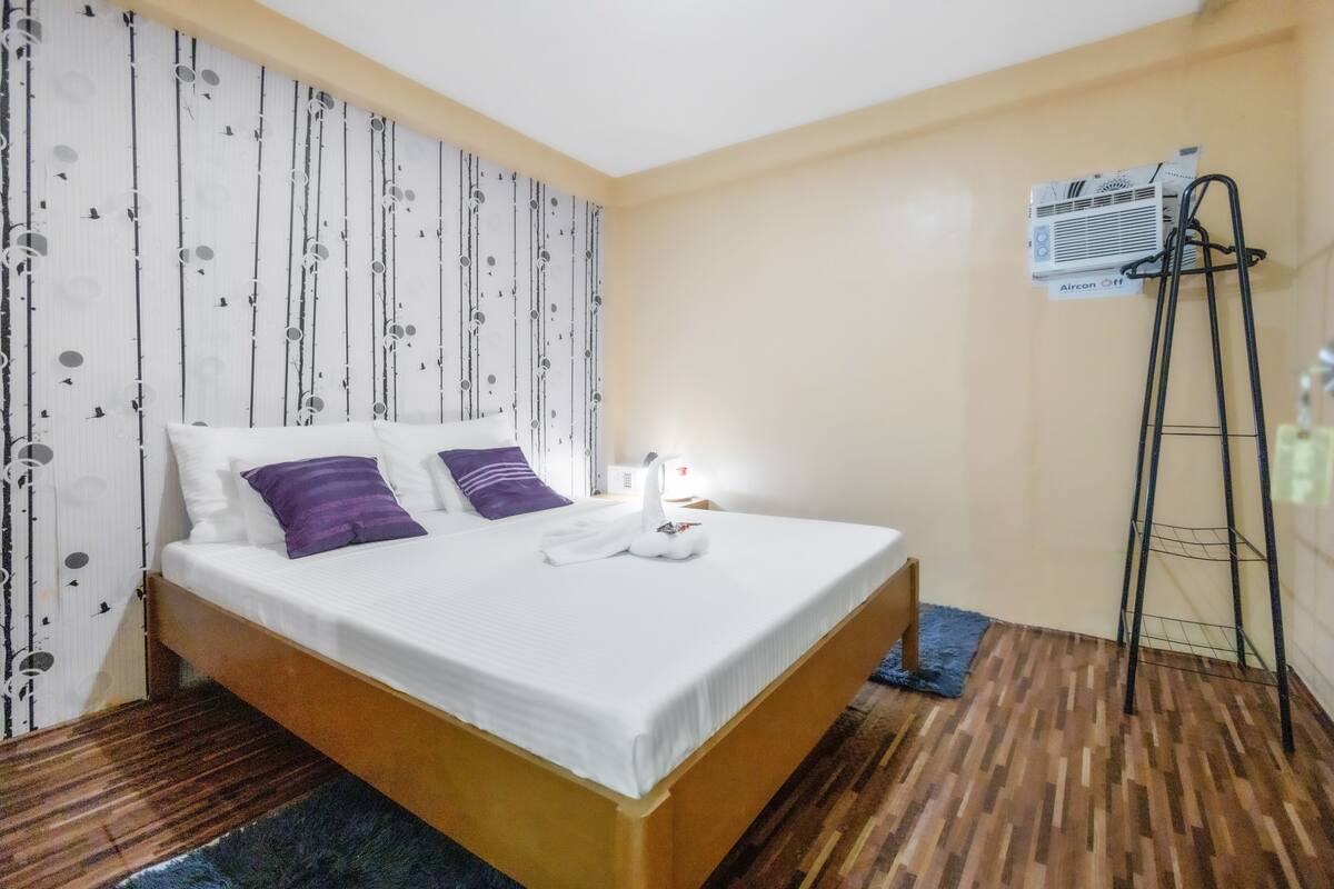 A neatly made bed with white and purple pillows, a wooden headboard, and a blue rug. There is a standing air conditioner on the wall and a black metal shelf.