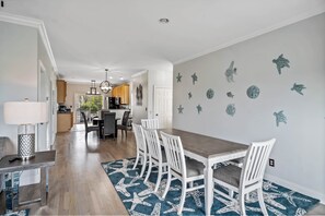 Uma sala de jantar com mesa de madeira e cadeiras brancas, um tapete azul estampado e uma cozinha ao fundo.