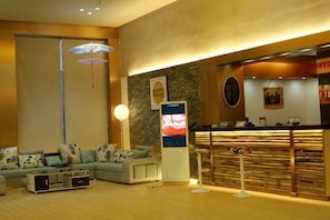 A reception area with a wooden reception desk, a seating area with a sofa and chairs, a standing fan, and a wall-mounted digital display.