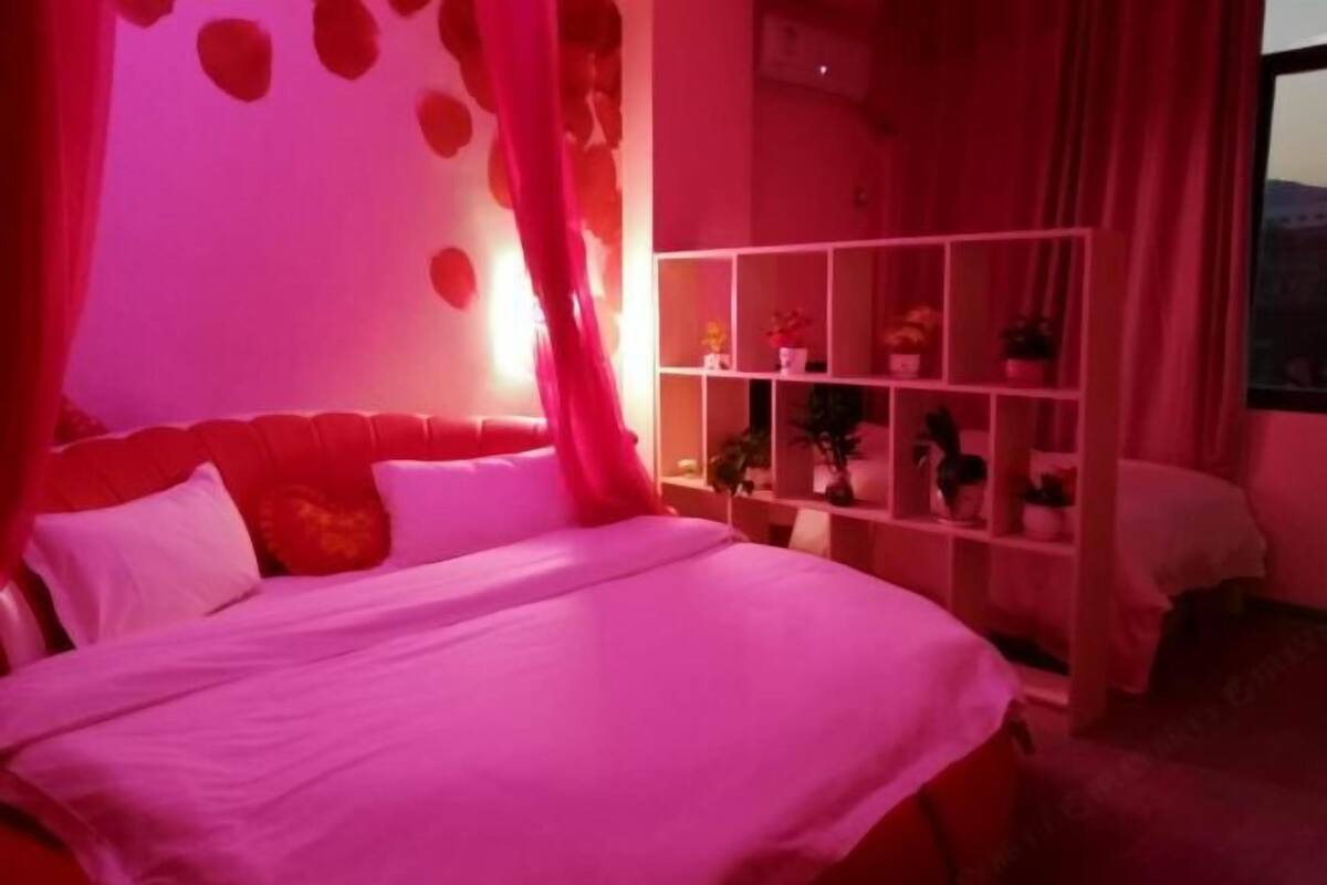 A bedroom with a bed, a white headboard, a red curtain, and a white shelf with various items.