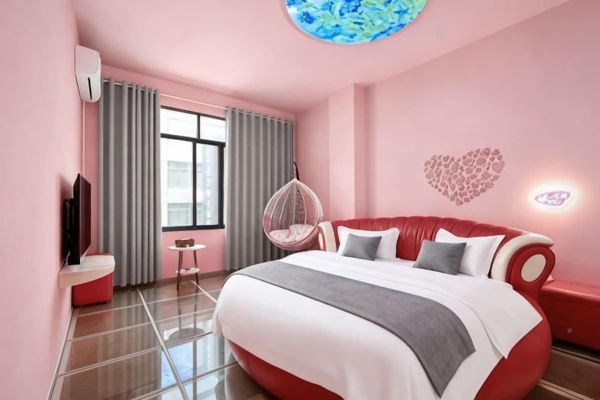 A pink-themed hotel room with a large bed, a TV, a small table, and a hanging chair.